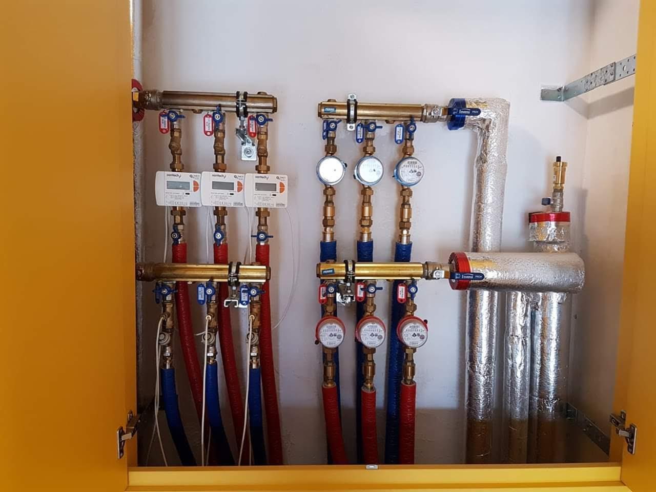 Rozdzielacz instalacji centralnego ogrzewania i wodnej z licznikami, rurami miedzianymi i plastikowymi, zabudowany w żółtej szafce, widoczne zawory i izolacja termiczna.