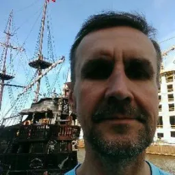 Człowiek robiący selfie na tle żaglowca i budynku w budowie. Ujęcie z dołu, niebieskie niebo w tle.