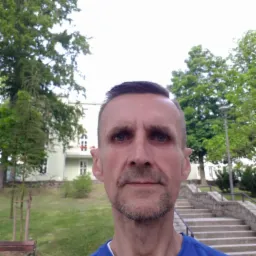 Portret mężczyzny w niebieskiej koszulce na tle parku z budynkiem i schodami.