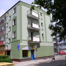 Fasada narożnego budynku mieszkalnego z balkonami, pomalowana w odcieniach zieleni, z niebieskimi drzwiami wejściowymi do lokalu usługowego na parterze i szyldem 'Obuwie Skórzane'; w tle inne...