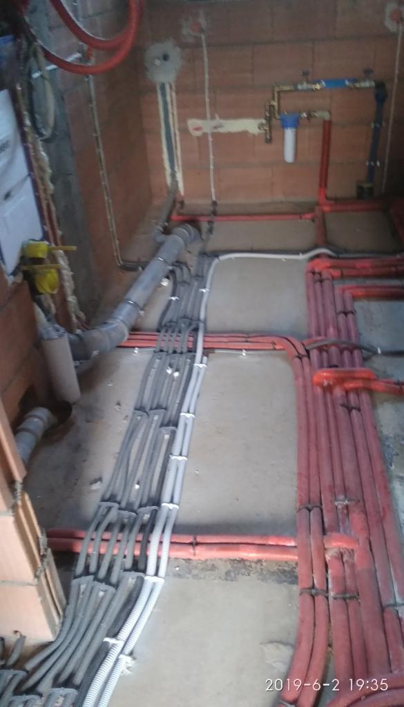 Surowe wnętrze w trakcie instalacji: liczne szare i czerwone rury oraz kable poprowadzone po podłodze i ścianach z cegły, widoczny filtr wody i fragment kanalizacji.