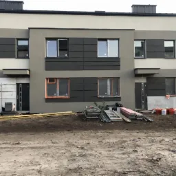 Trzysegmentowy budynek w trakcie prac elewacyjnych, z widocznymi rusztowaniami, zabezpieczonymi oknami i materiałami budowlanymi na ziemi.