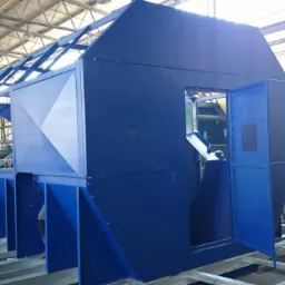 Niebieska, metalowa konstrukcja kabiny na ramie transportowej w hali produkcyjnej, widoczne elementy spawane i skręcane.