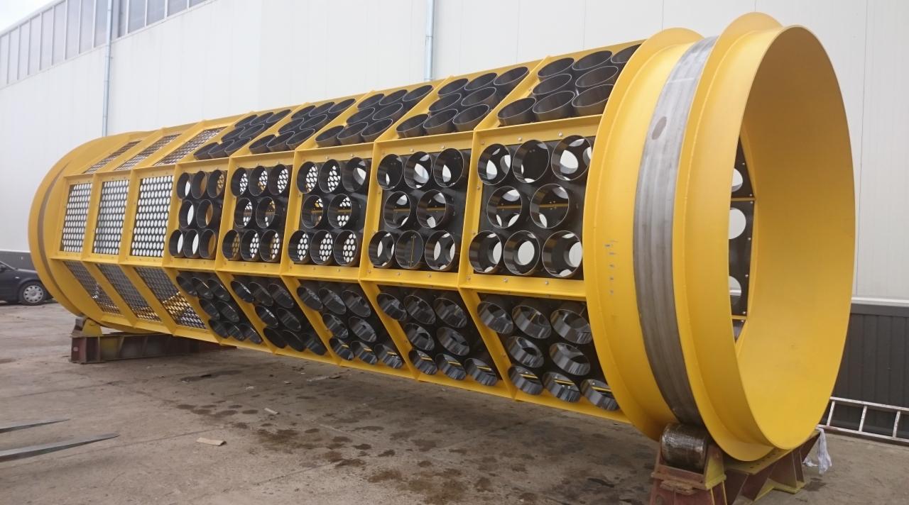 Żółta, cylindryczna konstrukcja z metalu, z licznymi otworami o okrągłym przekroju, umieszczona na metalowych podporach. Widoczne spawane elementy i siatka ochronna. Budynek przemysłowy w tle.