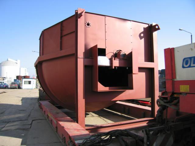 Duży, pomalowany na czerwono metalowy zbiornik o skomplikowanej konstrukcji, umieszczony na platformie transportowej. Widoczne elementy spawane i otwory montażowe. W tle budynek przemysłowy z kopułą.