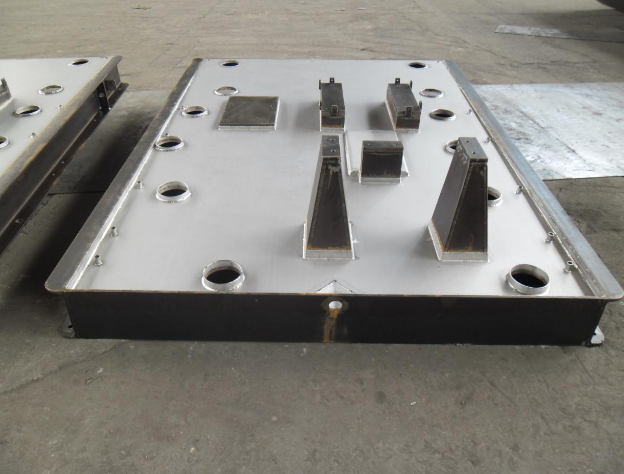 Duża, prostokątna, metalowa płyta z przyspawanymi elementami o różnych kształtach, leżąca na betonowej podłodze. Widoczne otwory i spawy.
