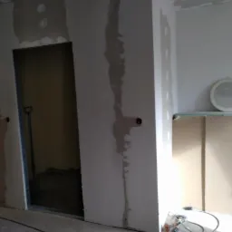 Remontowany pokój z widoczną ścianą w trakcie szpachlowania, otworem drzwiowym z widocznymi kablami w środku, oraz elementami instalacji elektrycznej i narzędziami na podłodze.