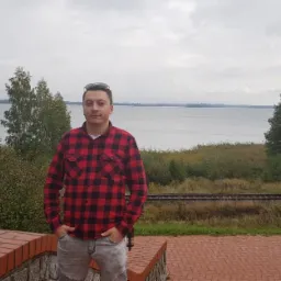 Mężczyzna w czerwono-czarnej koszuli w kratę stoi na tle jeziora z żaglówką, widoczne również tory kolejowe i butelka napoju na murku.