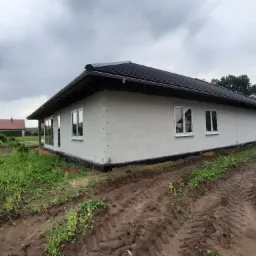 Budynek w trakcie budowy z zamontowanymi oknami PCV, widoczne ślady opon na ziemi i zachmurzone niebo.