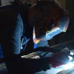 Pracownik w masce spawalniczej z motywem ognia łączy metalowe elementy, widoczny jasny łuk elektryczny i iskry.