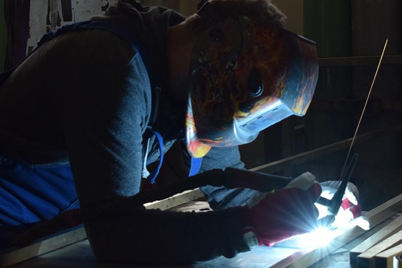 Pracownik w masce spawalniczej z motywem ognia łączy metalowe elementy, widoczny jasny łuk elektryczny i iskry.