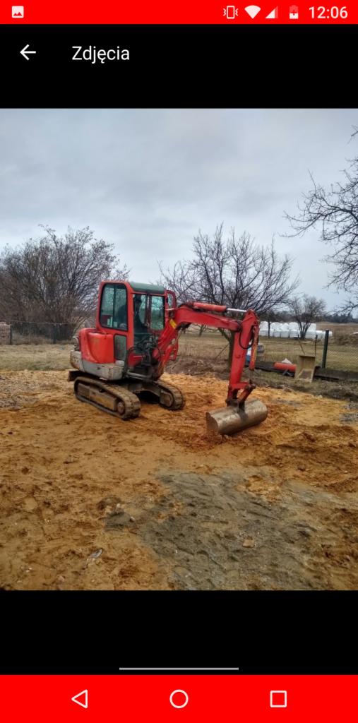 Czerwona mini koparka na górzystym terenie z piaskiem, widoczne drzewa bez liści w tle.