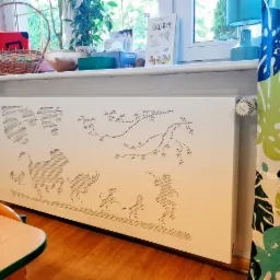 Biały panel grzejnikowy z dekoracyjnymi, perforowanymi wzorami zwierząt i roślin, umieszczony pod oknem z kolorowymi zasłonami w liście.