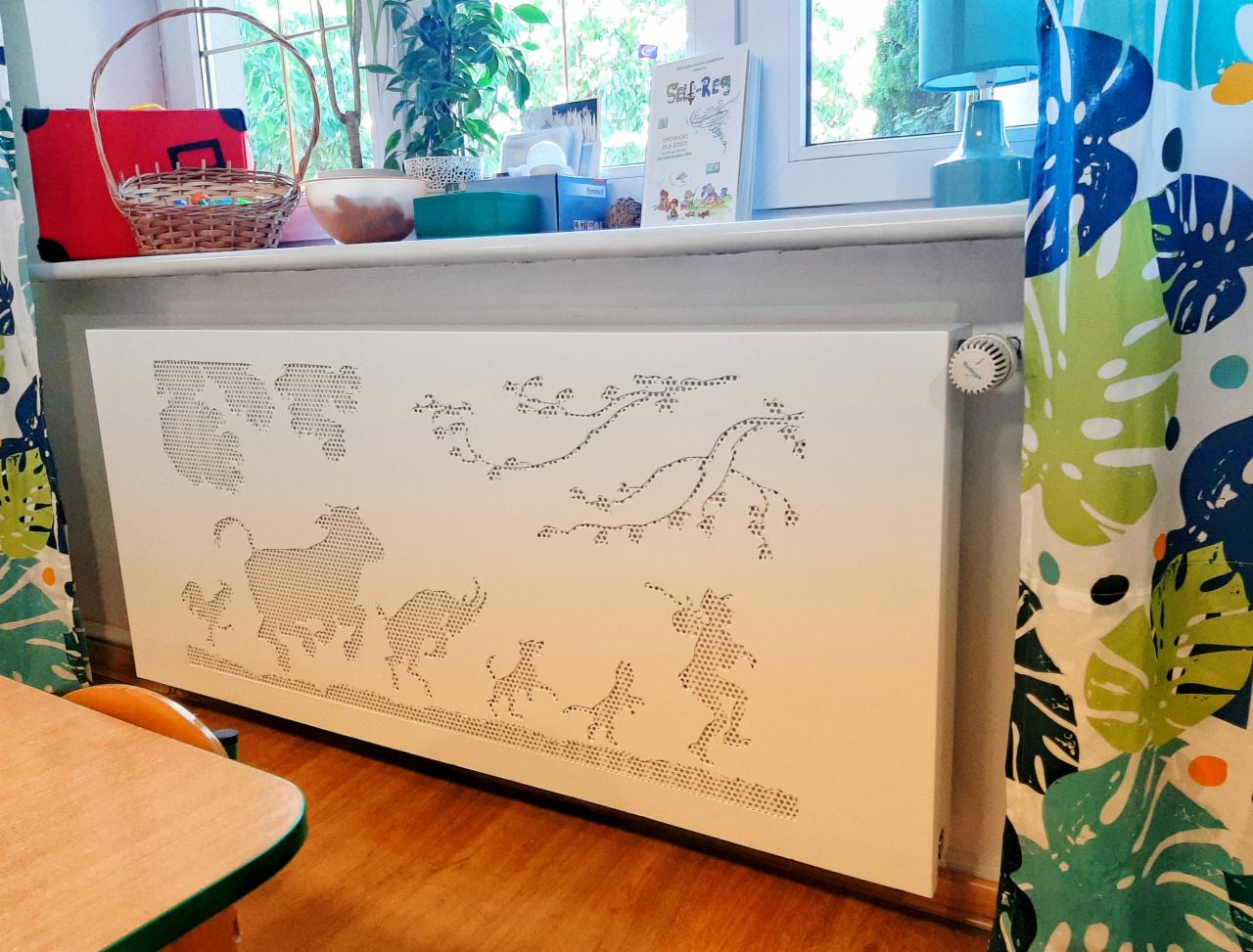 Biały panel grzejnikowy z dekoracyjnymi, perforowanymi wzorami zwierząt i roślin, umieszczony pod oknem z kolorowymi zasłonami w liście.