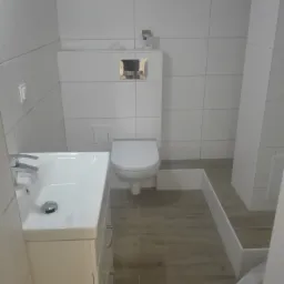 Nowoczesna, jasna łazienka po remoncie z białą umywalką z szafką, podwieszanym WC z chromowanym przyciskiem spłukiwania i podłogą imitującą drewno.