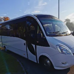 Biały bus pasażerski z przyciemnianymi szybami, zaparkowany na trawniku, z widocznym fragmentem jesiennego drzewa w tle.