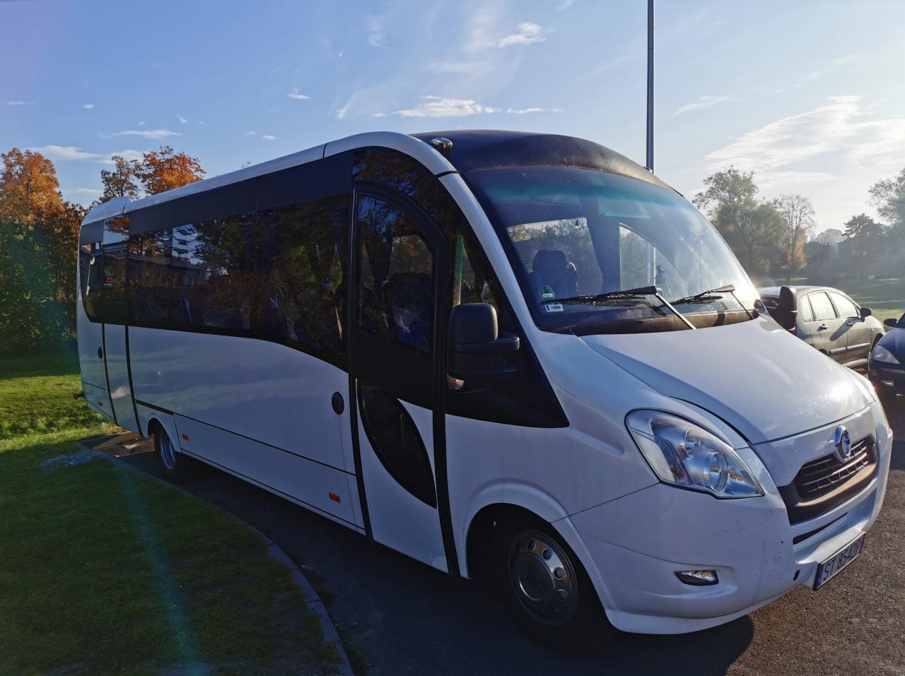 Biały bus pasażerski z przyciemnianymi szybami, zaparkowany na trawniku, z widocznym fragmentem jesiennego drzewa w tle.
