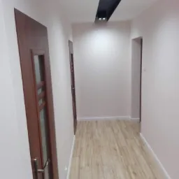 Korytarz po remoncie z drewnopodobną podłogą, pomalowanymi ścianami w odcieniach bieli i brązu oraz nowoczesnym oświetleniem sufitowym.