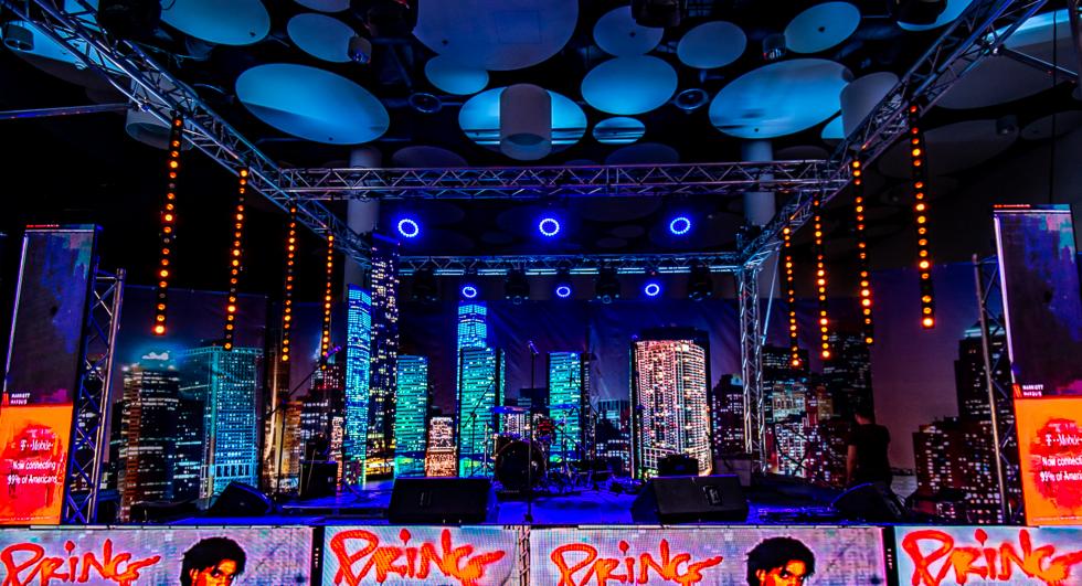 Scena z podświetlonym tłem imitującym panoramę miasta nocą, oświetlenie punktowe i dekoracje z motywem Prince'a na dolnych panelach, widoczny zestaw perkusyjny i elementy nagłośnienia.