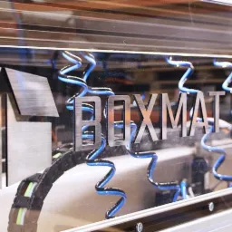 Zbliżenie na logo 'BOXMAT HD' wycięte z metalu, z niebieskimi przewodami spiralnymi w tle, zamontowane na przezroczystej obudowie urządzenia.