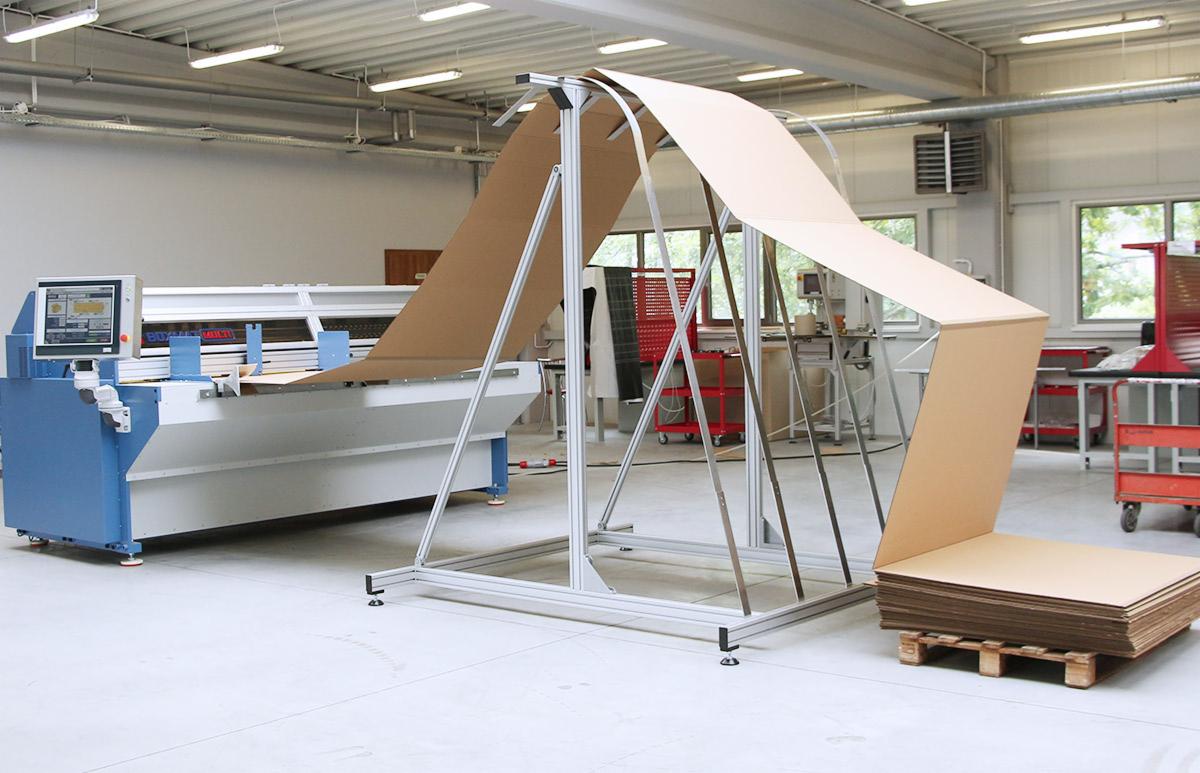 Maszyna do gięcia kartonu z ułożonymi arkuszami na palecie w tle hali produkcyjnej.