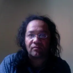 Portret mężczyzny z długimi, ciemnymi włosami i okularami w metalowych oprawkach, ubrany w niebieską koszulę w kratę, na rozmytym tle.