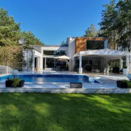 Nowoczesny dom z dużymi przeszkleniami, basenem i tarasem z białym zadaszeniem oraz huśtawką ogrodową, otoczony zielenią i wysokimi sosnami w słoneczny dzień.