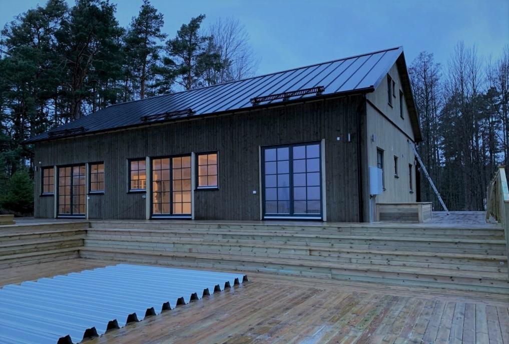 Nowoczesny dom z ciemnego drewna z metalowym dachem, oświetlony od wewnątrz, z dużym drewnianym tarasem i częściowo zakrytym basenem, otoczony drzewami.