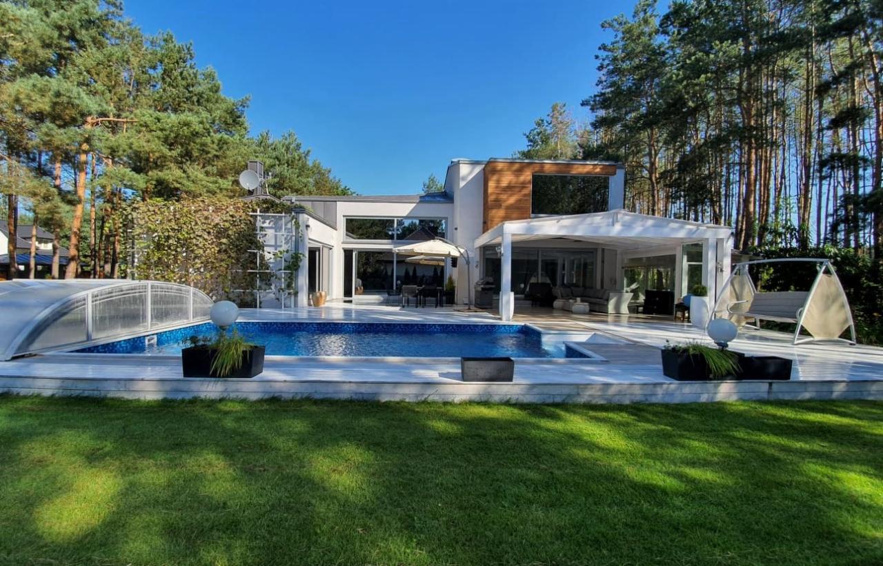 Nowoczesny dom z dużymi przeszkleniami, basenem i tarasem z białym zadaszeniem oraz huśtawką ogrodową, otoczony zielenią i wysokimi sosnami w słoneczny dzień.