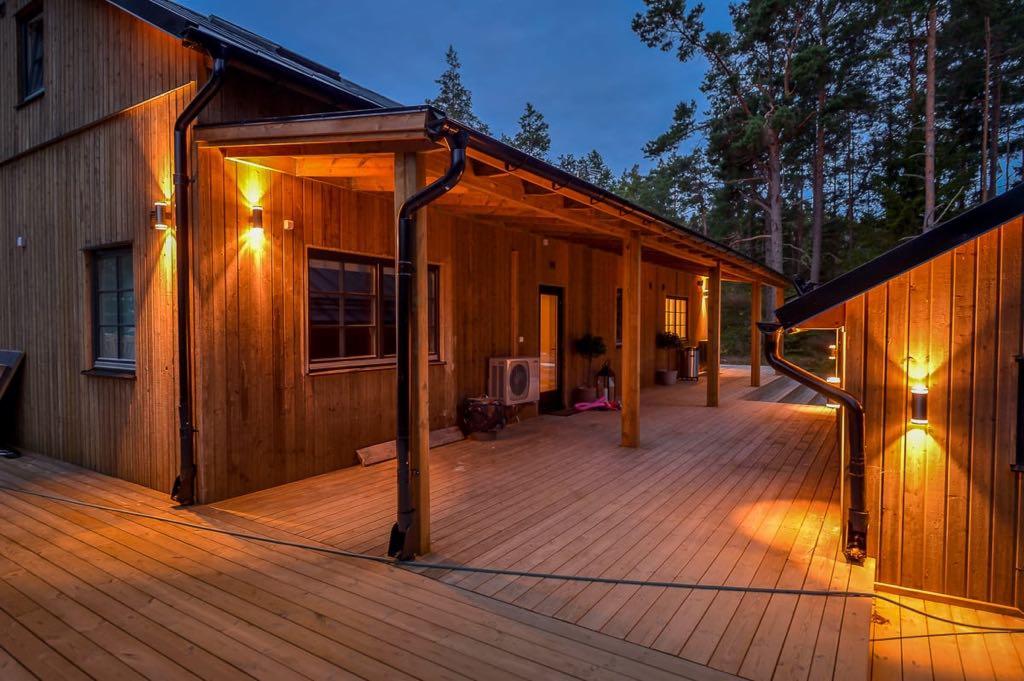 Oświetlony drewniany dom z tarasem w Skrzeszewie, widok z zewnątrz o zmierzchu, z widocznymi detalami konstrukcyjnymi i lampami ściennymi.