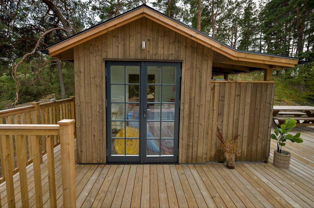 Mały drewniany domek z dwuskrzydłowymi przeszklonymi drzwiami, taras z desek, w tle drzewa
