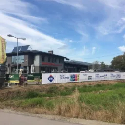 Budynek składu budowlanego Komplex-Pol z panelami słonecznymi na dachu, otoczony banerami reklamowymi klejów Atlas i kostką Libet, widoczne flagi reklamowe i góry w tle.