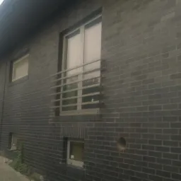 Elewacja domu z ciemnej, grafitowej cegły klinkierowej, widoczne okna z metalową balustradą oraz ścieżka z kostki brukowej prowadząca wzdłuż budynku.