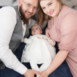 Rodzice z niemowlęciem w białej sukience leżący na ciemnoniebieskim tle, ojciec w kraciastej kamizelce i brązowej muszce, matka w pudrowej sukience, wszyscy uśmiechnięci.