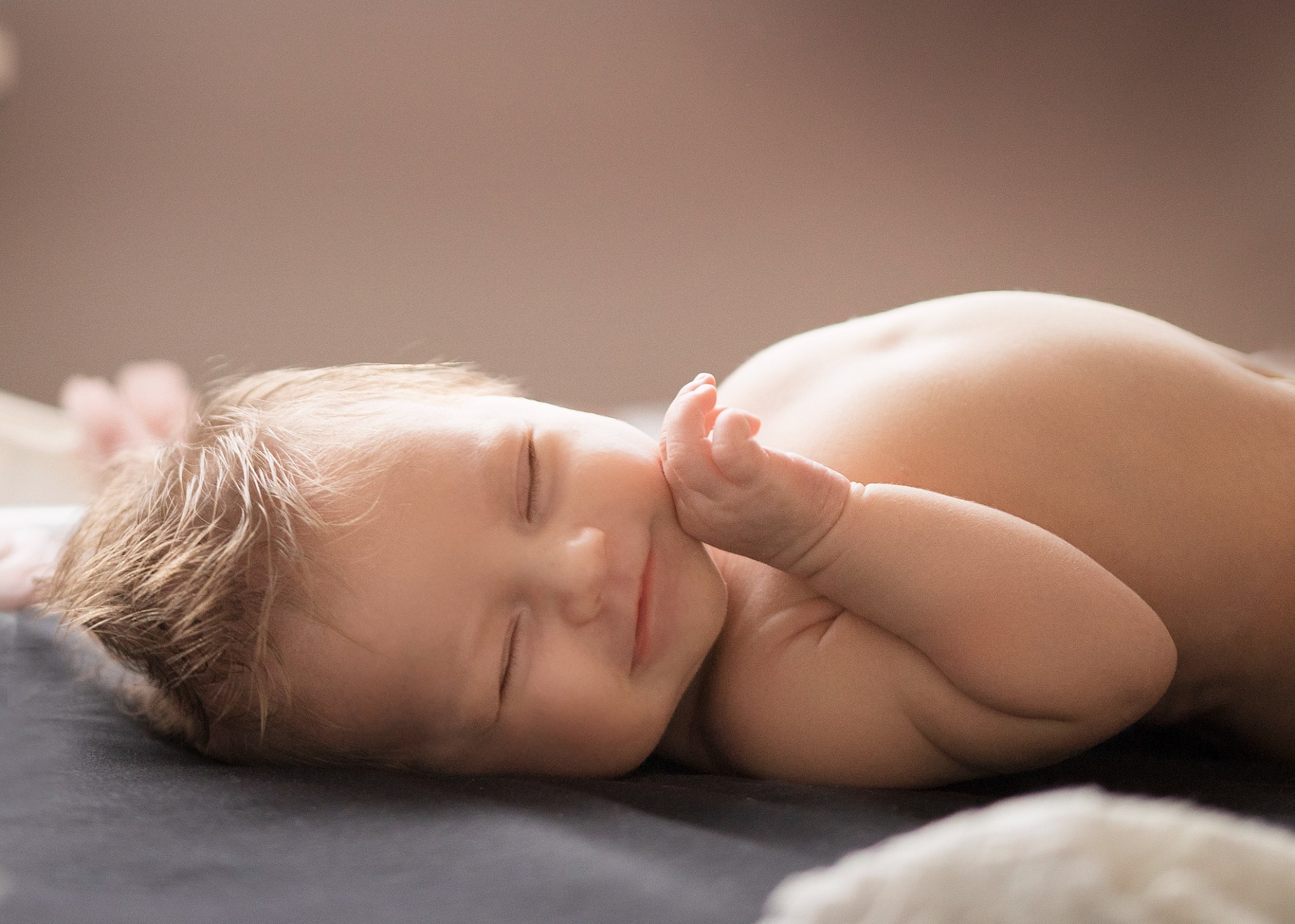 Śpiący noworodek leży na boku na ciemnoszarym kocyku, z delikatnym uśmiechem na twarzy i rączką pod policzkiem, w miękkim, naturalnym świetle.
