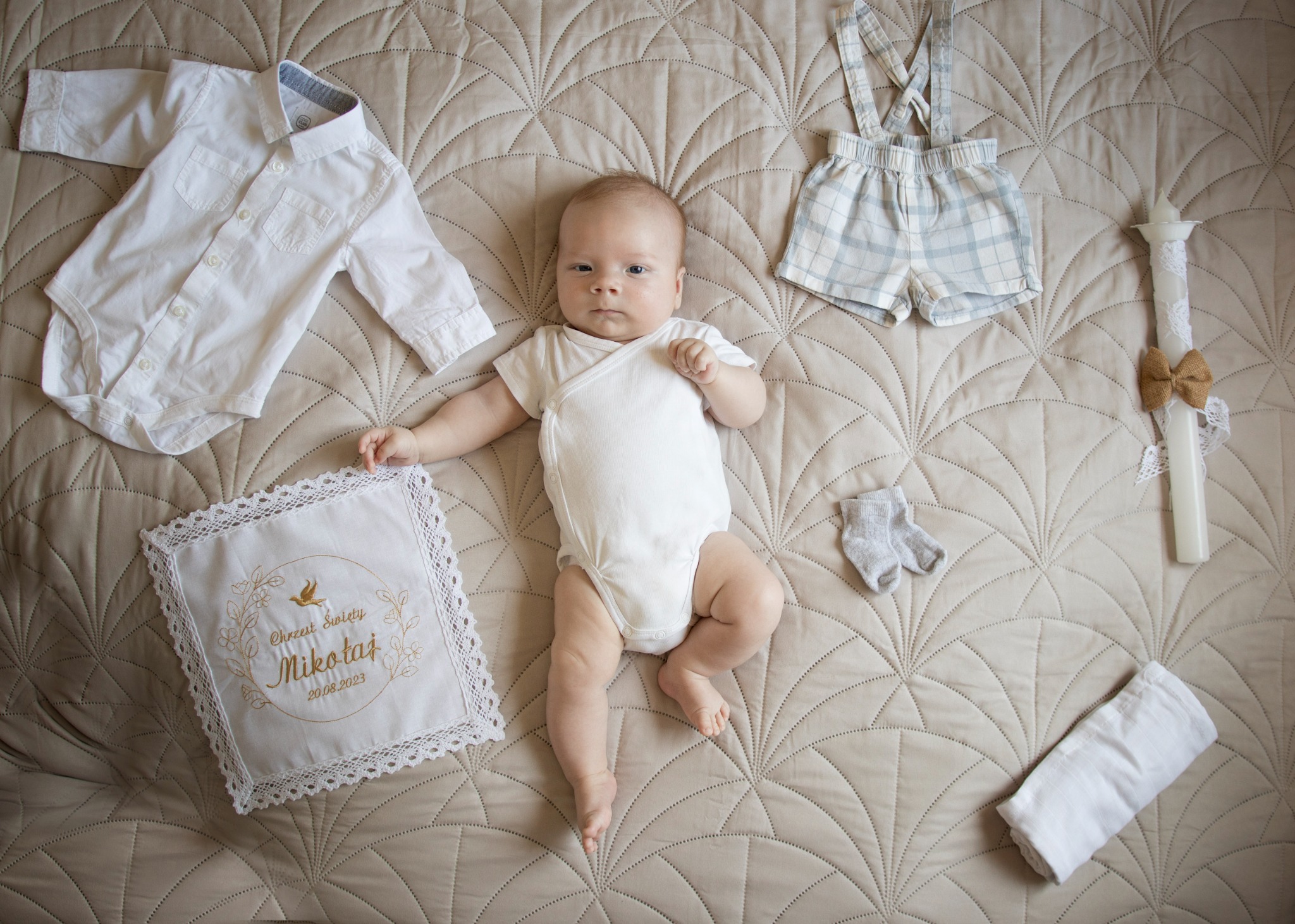 Niemowlę w białym body leży na beżowej kołdrze obok ubranka, świecy z kokardką i ozdobnej poduszki z napisem 'Chrzest Święty Mikołaj 20.08.2023'.