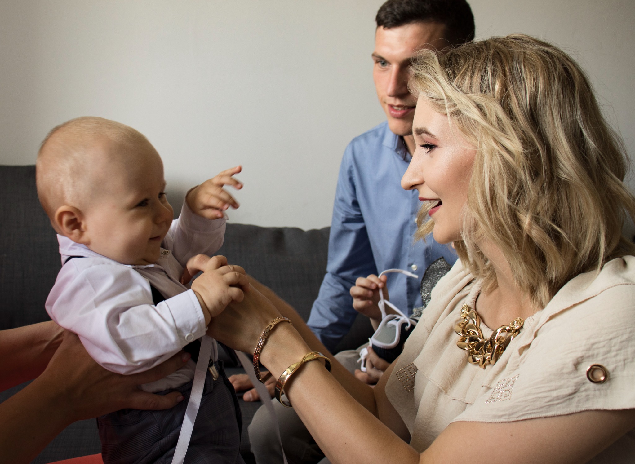 Uśmiechnięta blond kobieta w złotej biżuterii trzyma za ręce elegancko ubranego chłopca, w tle widoczny mężczyzna w niebieskiej koszuli trzymający buciki niemowlęce.
