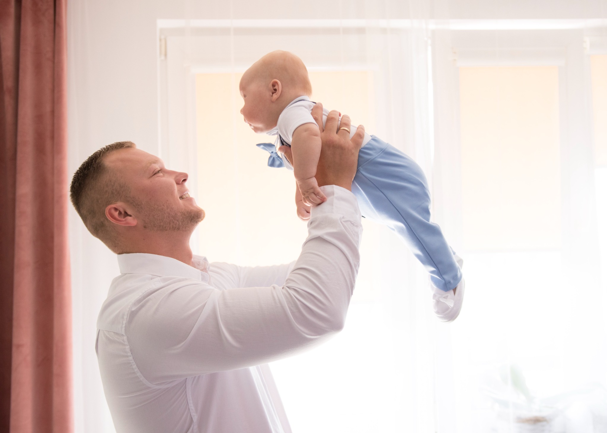 Uśmiechnięty tata trzyma wysoko w górze niemowlę ubrane w błękitny garniturek z muszką, jasne, naturalne światło z okna w tle.