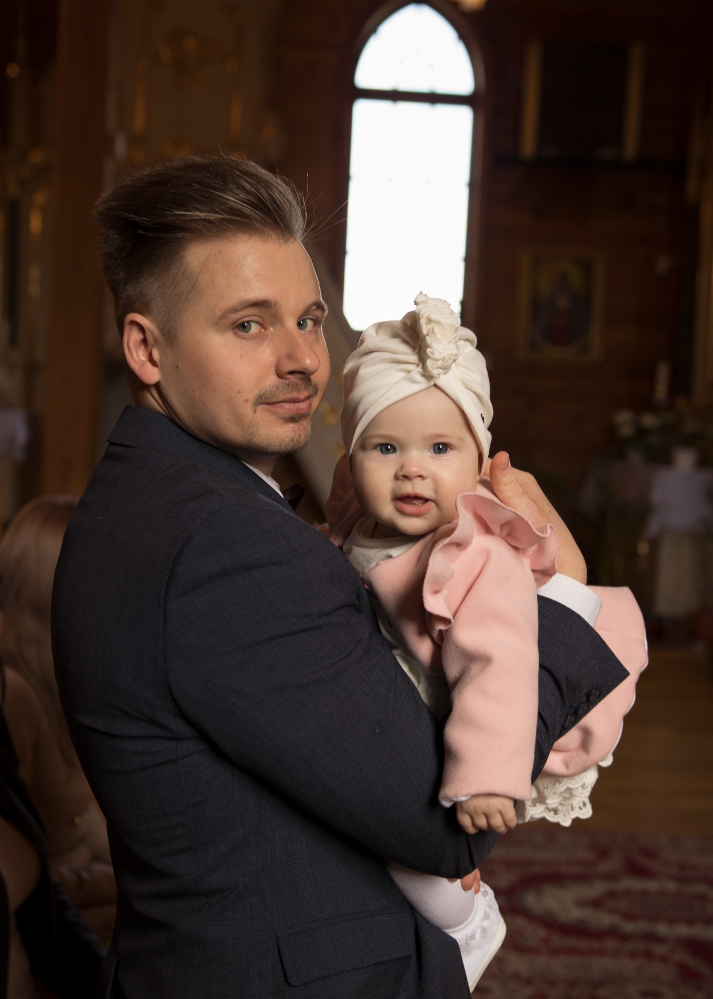 Mężczyzna w ciemnym garniturze trzyma na rękach uśmiechniętą dziewczynkę w różowym płaszczyku i białym turbanie na tle wnętrza kościoła.