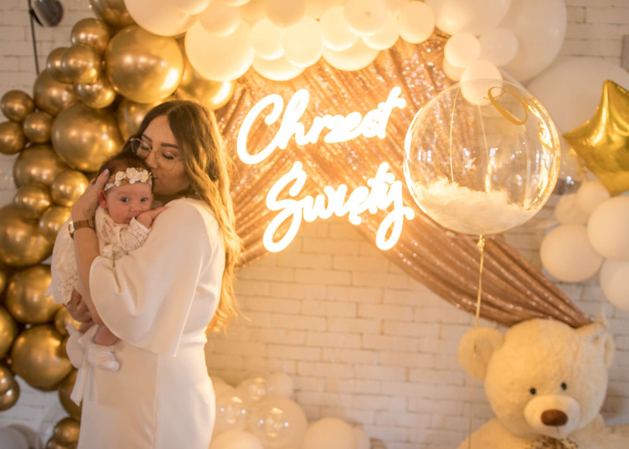 Mama całuje w czoło noworodka w Suwałkach, na tle dekoracji z białych i złotych balonów oraz napisu 'Chrzest Sweet'.