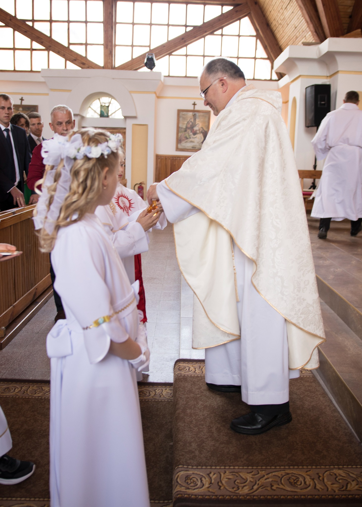 Dziewczynka w białej albie przyjmuje komunię świętą z rąk księdza w kościele, na głowie ma wianek z białych kwiatów.