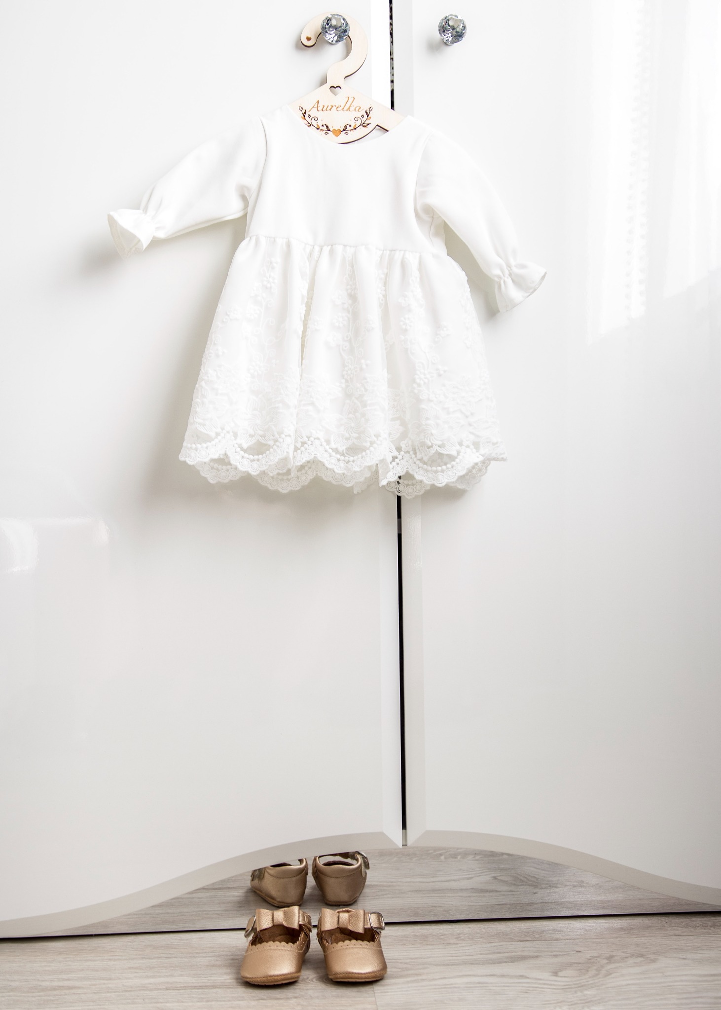 Biała, koronkowa sukienka niemowlęca zawieszona na drewnianym wieszaku z imieniem Aurelia, poniżej para złotych bucików na drewnianej podłodze, na tle białej szafy.