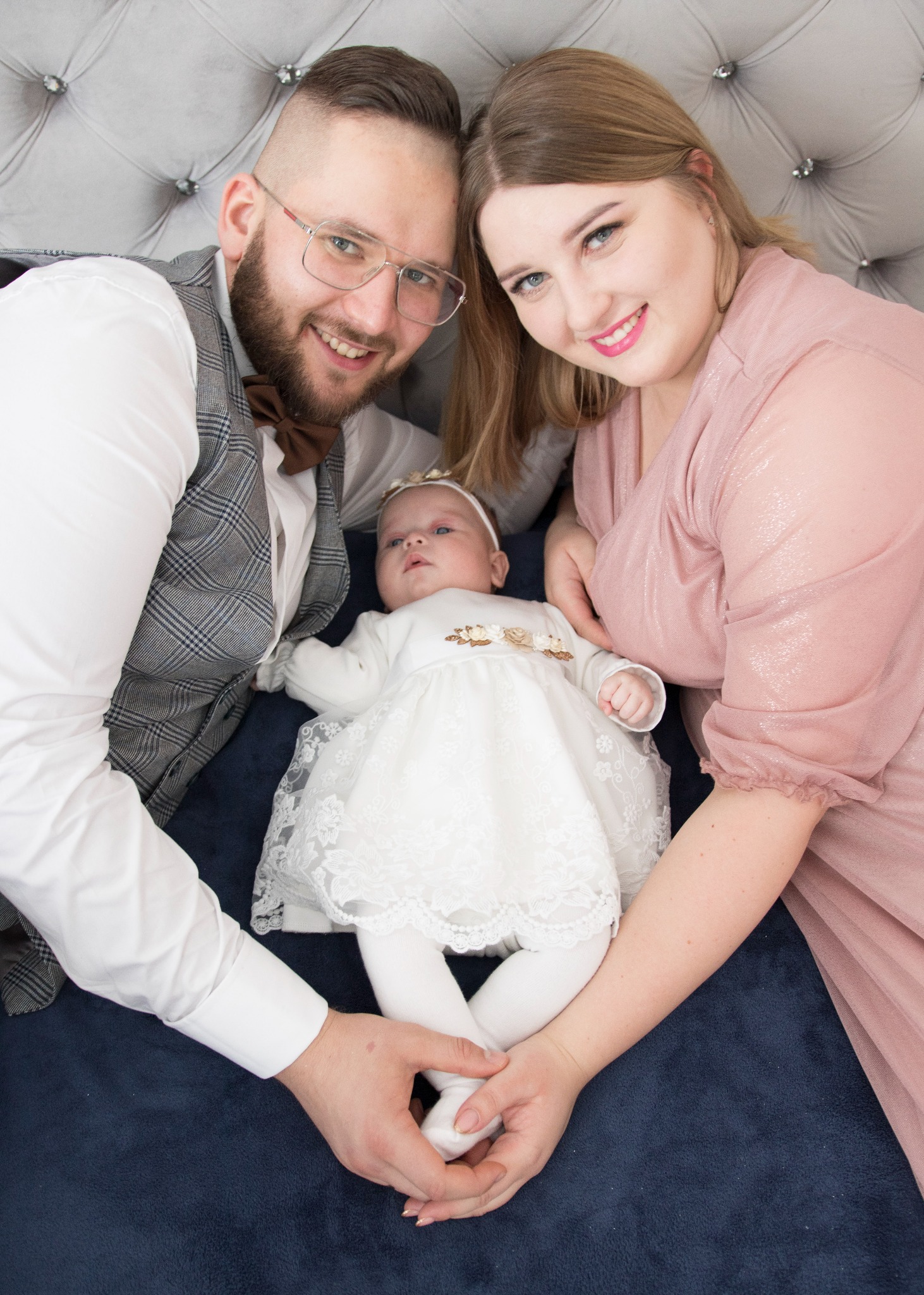 Rodzice z niemowlęciem w białej sukience leżący na ciemnoniebieskim tle, ojciec w kraciastej kamizelce i brązowej muszce, matka w pudrowej sukience, wszyscy uśmiechnięci.