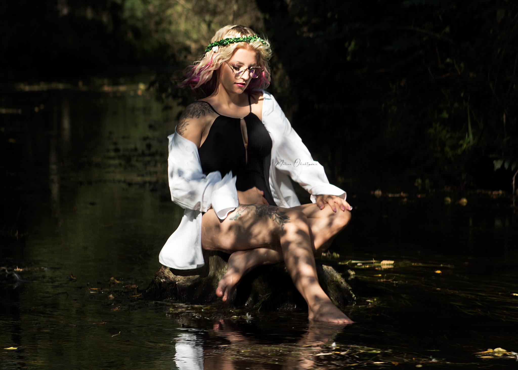 Kobieta w czarnym stroju kąpielowym i białej koszuli siedzi na pniu drzewa w rzece, z wiankiem z kwiatów na głowie i okularami, delikatne światło pada na jej twarz i ciało, widoczne tatuaże...