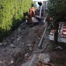 Pomarańczowa mini koparka w trakcie prac ziemnych przy wykopie w ogrodzie, widoczne fragmenty gruzu i narzędzia.