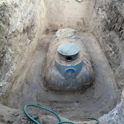 GREEN-BAU TECHNIK Cezary Palęga - Montaż szamba betonowego w wykopie, widoczne narzędzia i zielony wąż, ściany wykopu z warstwami ziemi.