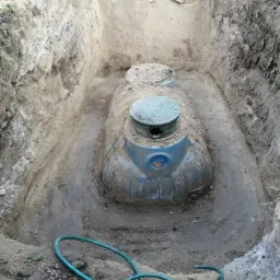 Montaż szamba betonowego w wykopie, widoczne narzędzia i zielony wąż, ściany wykopu z warstwami ziemi.