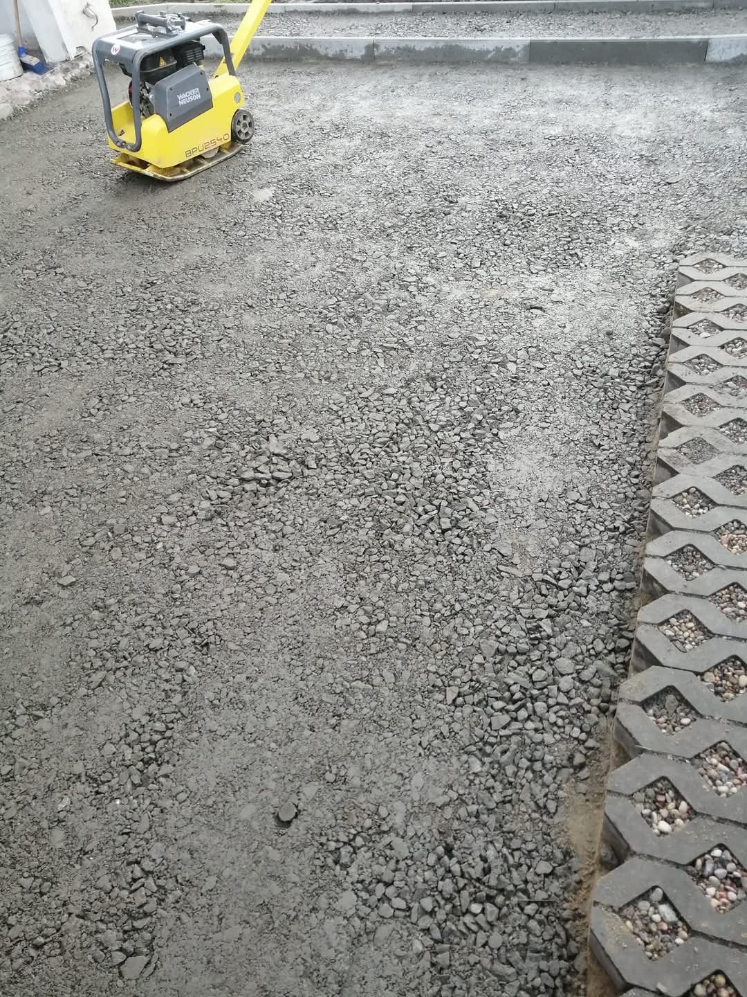 Żółta zagęszczarka Wacker Neuson ubija szarą nawierzchnię z kruszywa obok ażurowych płyt betonowych wypełnionych kamieniami.