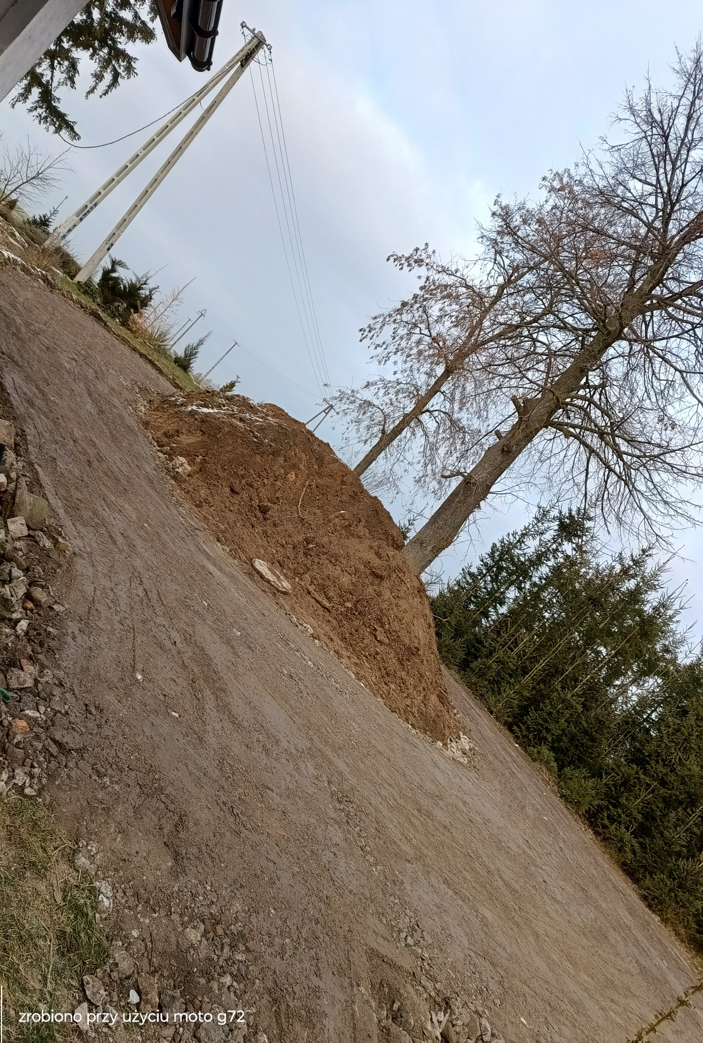 Skarpa ziemna z drzewami na zboczu, widoczne słupy energetyczne w tle, pochmurne niebo.