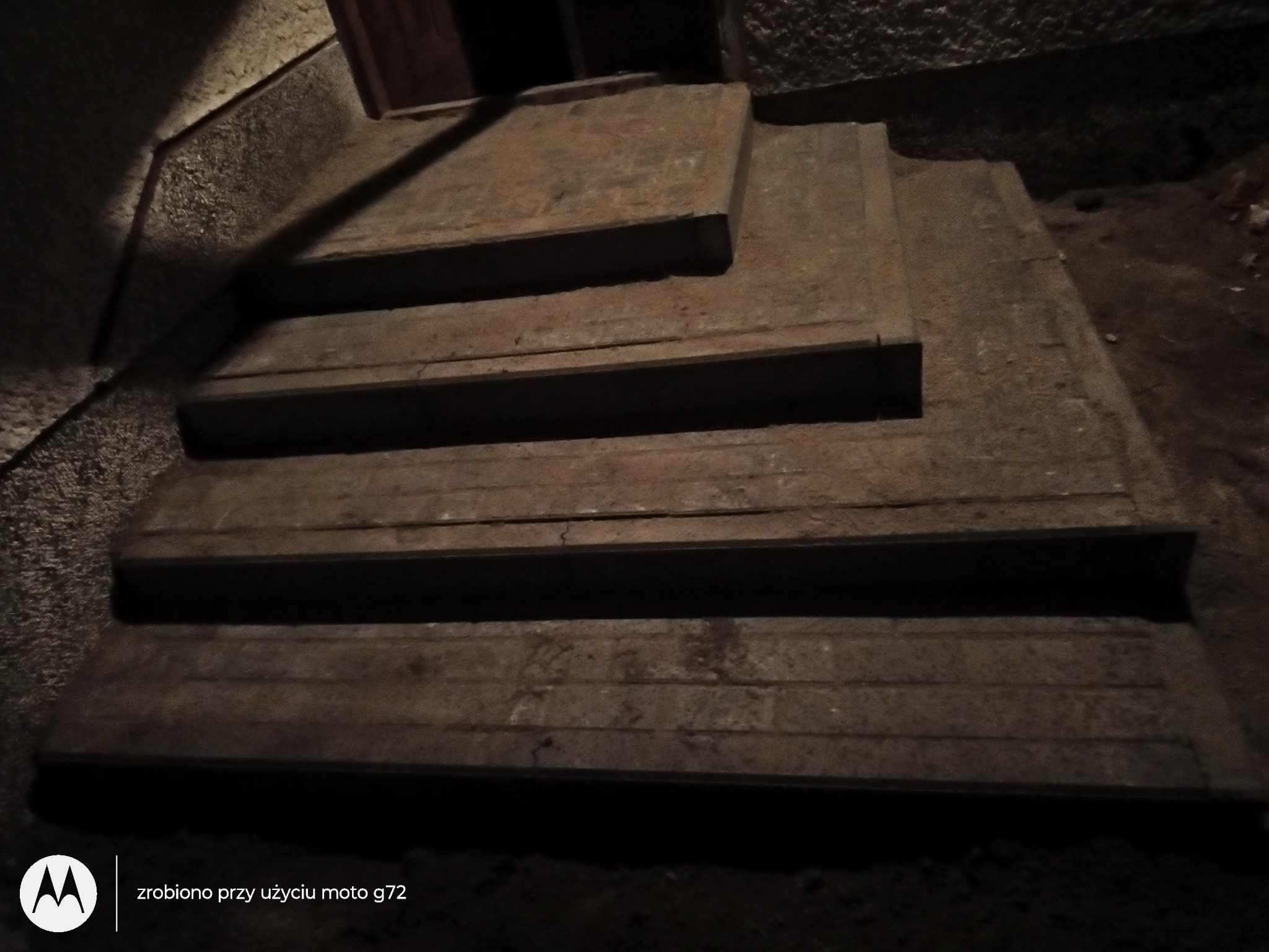 Betonowe schody zewnętrzne w słabym oświetleniu, prowadzące do ciemnego wejścia, widoczne ślady zużycia i zabrudzenia.